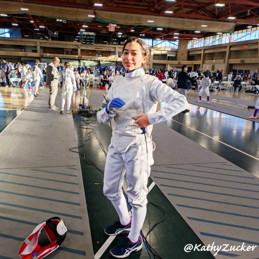 Young girl wearing fencing equipment stands holding wire attached to piste at large tournament