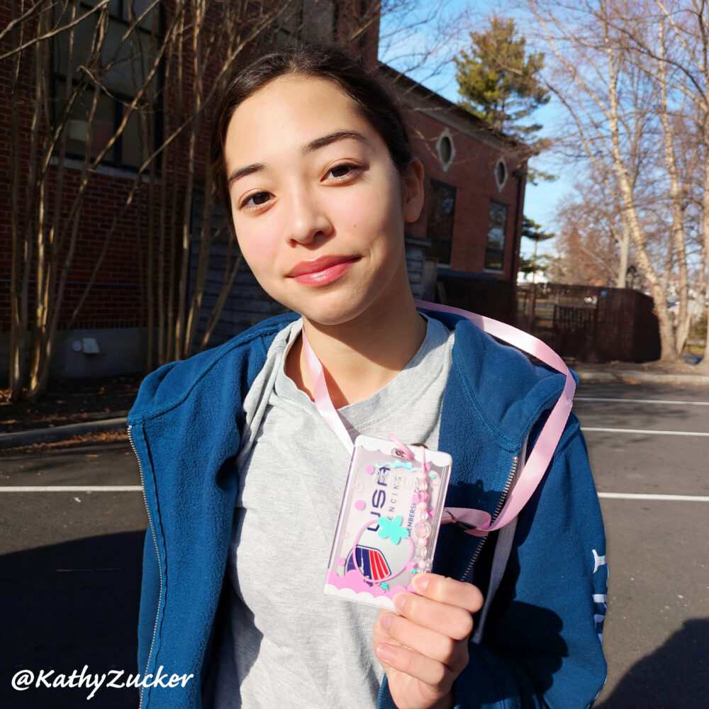 A young girl wearing a USA Fencing card in a pink badge holder stands in a parking lot