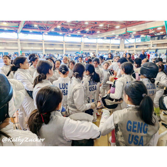 Young girls wearing fencing equipment wait for direct elimination strip assignments at a large tournament