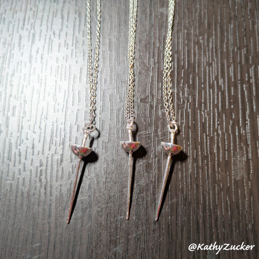 Three sterling silver fencing épée weapon pendants hang suspended from chains on a wood grain surface