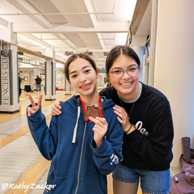 Two sisters wearing sweatshirts display a medal while standing in a fencing club